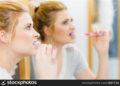Woman brushing cleaning teeth closeup. Blonde girl with toothbrush in bathroom, looking at mirror. Oral hygiene.. Woman brushing cleaning teeth in bathroom
