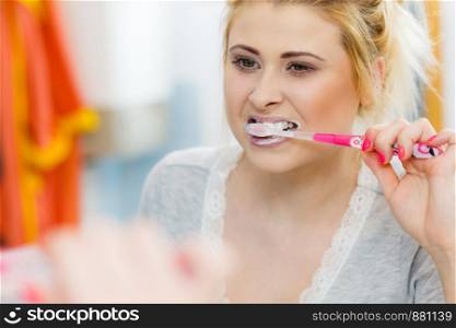 Woman brushing cleaning teeth closeup. Blonde girl with toothbrush in bathroom, looking at mirror. Oral hygiene.. Woman brushing cleaning teeth in bathroom