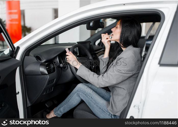 Woman buying new car in showroom, lady paints her lips with lipstick behind the wheel. Female customers choosing vehicle in dealership, automobile sale, auto purchase
