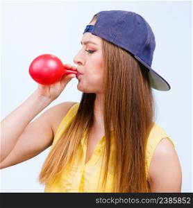 Woman casual style teenage hipster girl blowing inflating a red balloon, young female wearing cap having fun, studio shot on blue.. Woman casual style blowing up a red balloon