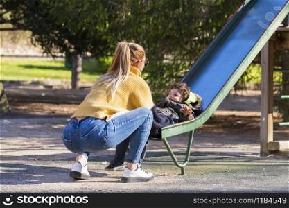 Woman catches adorable laughing child at the end of a metal slide in a playground on an out of focus background. Family and fun concept.. Woman catching boy at the end of a slide