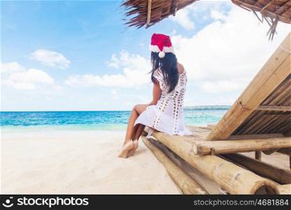 Woman celebrate Christmas on beach. Young woman in santa claus hat celebrating Christmas sitting in chaise lounge on tropical sea beach