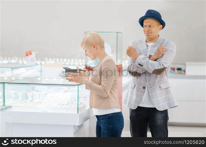 Woman choosing jewels, man in shock from the price, jewelry store. Love couple buying gold decoration in jewellery shop. Woman choosing jewels, man in shock from the price