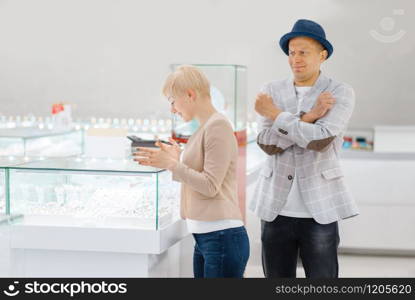 Woman choosing jewels, man in shock from the price, jewelry store. Love couple buying gold decoration in jewellery shop. Woman choosing jewels, man in shock from the price