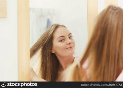 Woman combing brushing her long smooth hair in bathroom, looking in mirror. Girl taking care refreshing her hairstyle. Haircare concept.