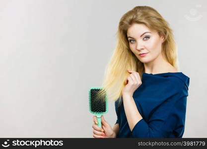 Woman combing her hair with brush. Young female with beautiful natural blond straight long hairs, studio shot on grey. Woman brushing her long hair with brush