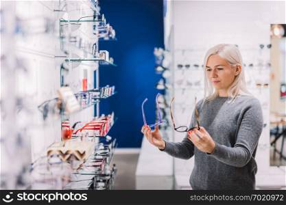 Woman comparing glasses at optic store. Eyewear shopping, correction glasses.. Woman comparing glasses at optic store.