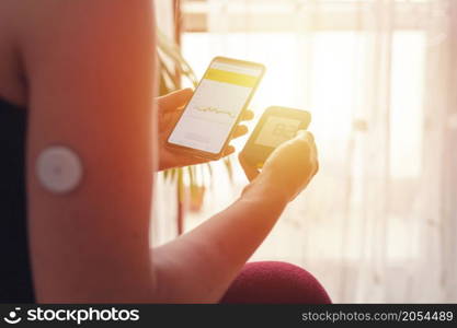 Woman comparing result of glucose level check on the traditional glucometer and on the mobile phone reading from modern technology remote sensor mounted on her forehand