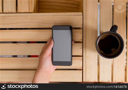 woman computer smartphone drink mug office supplies technological devices. Young lady holding a dark smartphone with left hand in a crate table and a mug of black coffee. Office supplies, cell phone, technological devices and wooden desk.