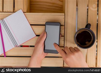 woman computer smartphone drink mug office supplies technological devices. Young lady using and holding a dark smartphone in a crate table with a mug of black coffee. Office supplies, cell phone, technological devices and wooden desk.