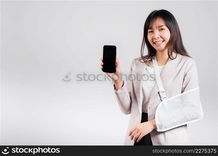 Woman confident smiling broken arm after accident and wear arm splint for treatment and hold smartphone, happy Asian female sling supported hand isolated on white background, social media, copy space