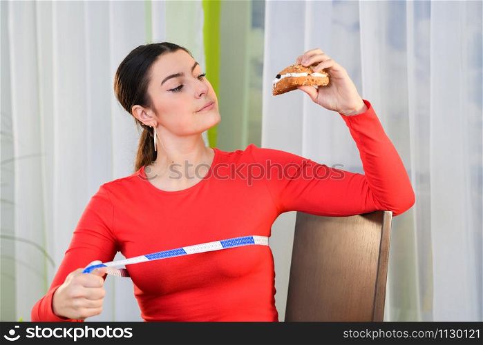 Woman considers eating a sandwich while using a measuring tape on an out of focus background. Healthy lifestyle concept.. Woman considers taking a bite of a sandwich