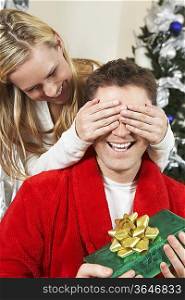 Woman covering eyes of man holding present by Christmas tree