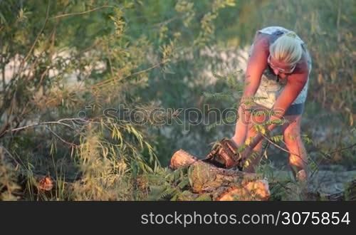 Woman cutting a log of dry wood on the ground into sections with chainsaw during weekend