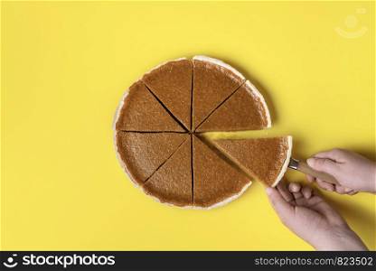 Woman cutting and taking a slice of pumpkin pie on a yellow paper background. Flat lay of traditional American pie. Thanksgiving sweet pastry item.