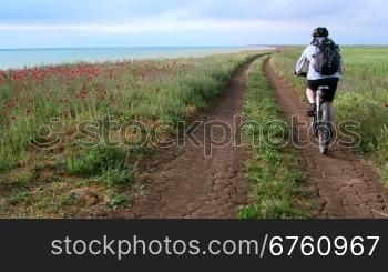 Woman cyclist rides bike along the seashore down a dirt road, rear view