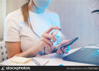 Woman disinfects the surface of the phone by sanitizer spray on the working place. Coronavirus concept. Woman in quarantine for coronavirus covid-19 working from home.. Woman disinfects the surface of the phone