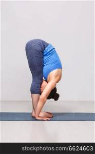 Woman doing yoga asana Uttanasana - standing forward bend pose on yoga mat in studio on grey bagckground. Woman doing yoga asana Uttanasana - standing forward bend