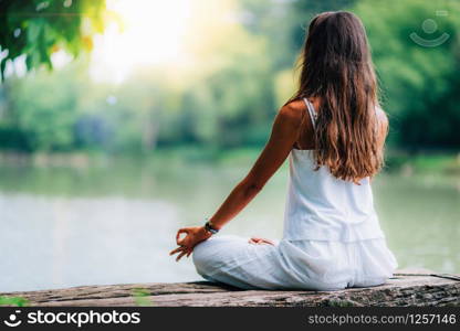 Woman doing yoga by the lake, sitting in lotus pose with fingers in mudra position.