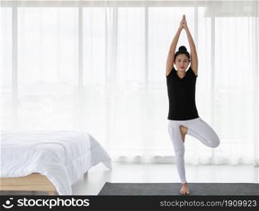 Woman doing yoga exercise at home, stretching in Tree pose or Vrikshasana svanasana in white bedroom after wake up in the morning. Healthcare concept.