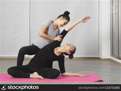 Woman doing yoga practice with private teacher at home class, working out with female yoga instructor. Trainer helps student to do Sukhasana pose or Easy seat pose, stretching with side bend. Personal yoga trainer and healthy lifestyle and fitness concept.