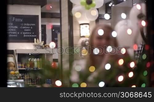 Woman drinking tea in cafe and looking at Christmas lights. View through twinkling garland. Defocus at the beginning.