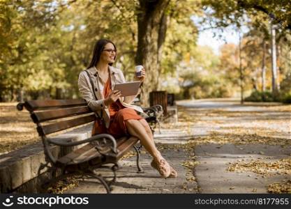 Woman drinks coffee and  sitting on bench in park during autumn weather, using tablet pc and  checking social media.