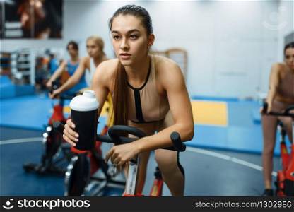 Woman drinks water on a stationary bike in gym. People on fitness workout in sport club, athletic girls in sportswear on training indoors. Woman drinks water on a stationary bike in gym