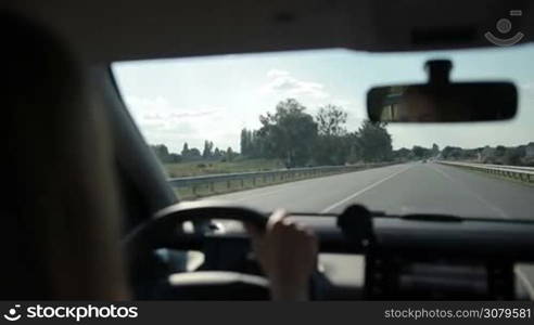 Woman driving car on motorway with rural scene on background. View from inside of vehicle&acute;s interior. Female driver controlling auto on high speed on freeway during sunny summer day.