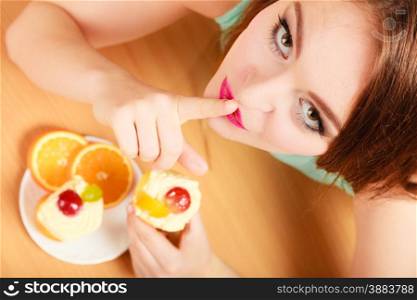 Woman eating delicious gourmet sweet cream cake cupcake and orange showing hand quiet sign gesture. Glutton girl having breakfast in secrecy. Appetite and gluttony concept.. Woman eating cake showing quiet sign. Gluttony.