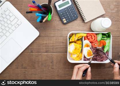 Woman eating healthy breakfast in box with rice berry, boiled eg. Woman eating healthy breakfast in box with rice berry, boiled egg, pumpkin, sweetcorn and tomatoes on workspace desk, Top view