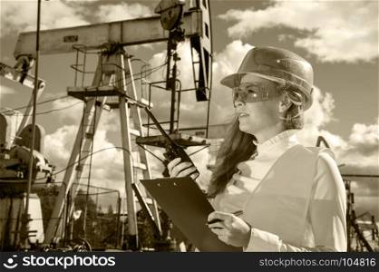 Woman engineer in the oil field talking on the radio wearing helmet and work clothes. Industrial site background. Oil and gas concept. Toned sepia.