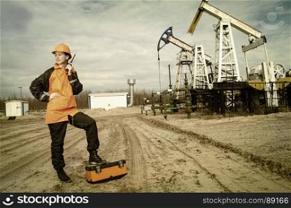 Woman engineer in the oil field talking on the radio wearing orange helmet and work clothes. Industrial site background. Toned.. Oil and gas industry worker.