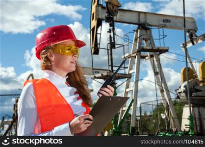 Woman engineer in the oil field talking on the radio wearing red helmet and orange work clothes. Industrial site background. Oil and gas concept.