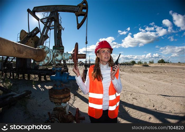 Woman engineer in the oil field talking on the radio wearing red helmet and orange work clothes. Industrial site background.