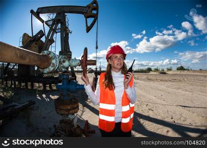Woman engineer in the oil field talking on the radio wearing red helmet and orange work clothes. Industrial site background.