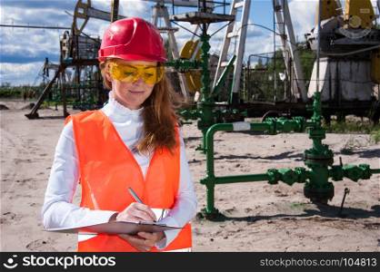 Woman engineer in yellow glasses on the oil field wearing red helmet and work clothes. Industrial site background.
