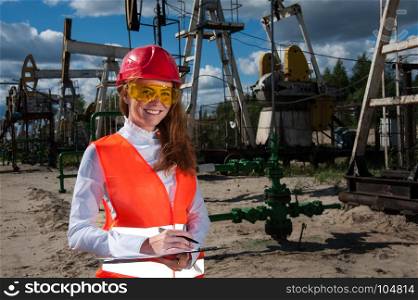 Woman engineer in yellow glasses on the oil field wearing red helmet and work clothes. Industrial site background.