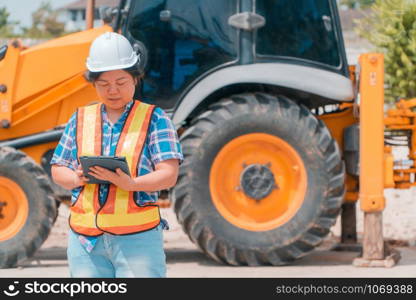 Woman Engineering wearing a white safety helmet standing In front of the backhoe And are using tablet to check the blueprint with construction