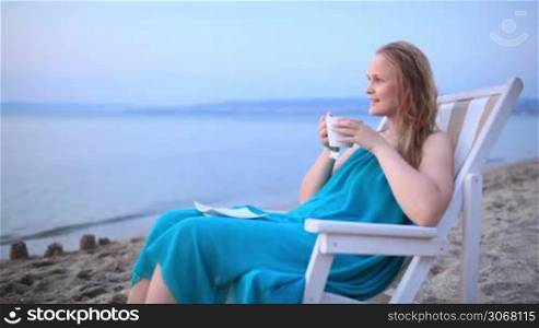 Woman enjoying a cup of tea at the seaside sitting relaxing on a deckchair with a blissful expression overlooking a tropical beach