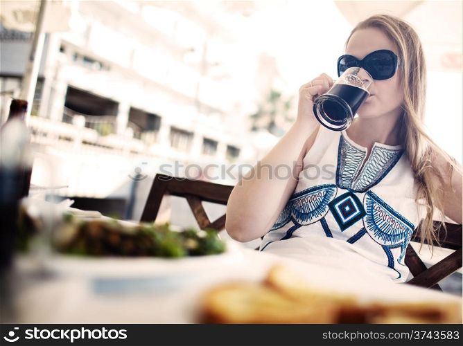 Woman enjoying a dark beer with her meal as she sits at a table at an open-air restaurant wearing a pair of sunglasses