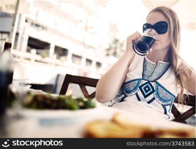 Woman enjoying a dark beer with her meal as she sits at a table at an open-air restaurant wearing a pair of sunglasses