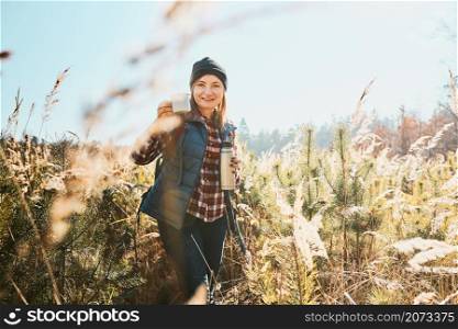 Woman enjoying the coffee in bright warm sunlight during vacation trip. Woman standing in tall grass and looking at camera holding cup of coffee and thermos flask. Woman with backpack hiking along path in mountains. Active leisure time close to nature