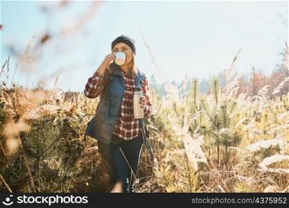 Woman enjoying the coffee in bright warm sunlight during vacation trip. Woman standing in tall grass and looking away holding cup of coffee and thermos flask. Woman with backpack hiking along path in mountains. Active leisure time close to nature