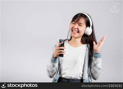 Woman excited smiling listening to music radio in bluetooth headphones and singing song on smartphone studio shot isolated white background, happy Asian young female Karaoke online app in mobile phone