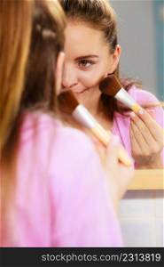 Woman face painting. Girl applying rouge or bronzing powder with brush to her skin looking in bathroom mirror. Makeup cosmetics and beauty procedures.. Woman applying bronzing powder with brush to her skin