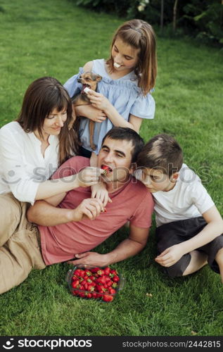 woman feeding strawberry husband while sitting with her children grass