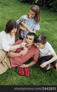 woman feeding strawberry husband while sitting with her children grass