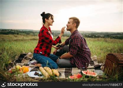 Woman feeds grapes to her man on picnic in summer field. Romantic junket of man and woman, love couple happy together. Woman feeds grapes to her man on picnic