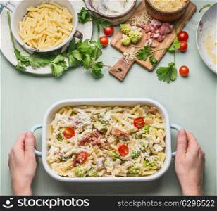 Woman female hands holding pasta casserole with romanesco cabbage and ham in creamy sauce, on kitchen table background with ingredients, top view, border. Italian cuisine